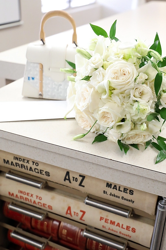 Bridal bouquet of white roses and blooms with greenery accents beside a small handbag on a countertop in soft window light