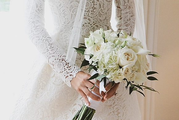 Bridal bouquet of white roses and hydrangea with greenery, ribbon wrap, held by bride in lace sleeves and veil in soft indoor light
