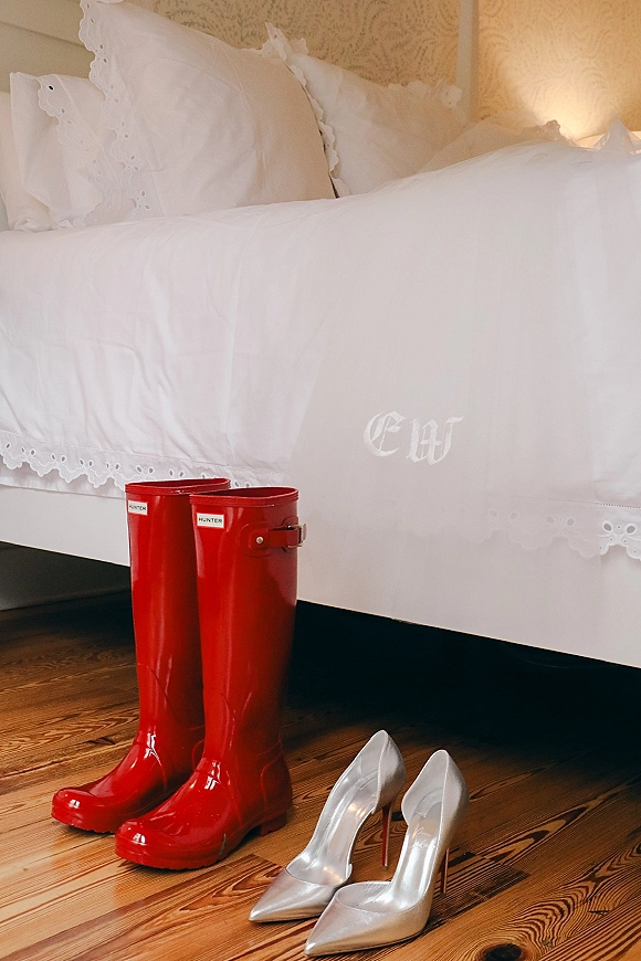 Bridal shoes silver wedding heels beside red rain boots on a wooden floor by a bed with monogrammed white duvet and ruffled pillows.