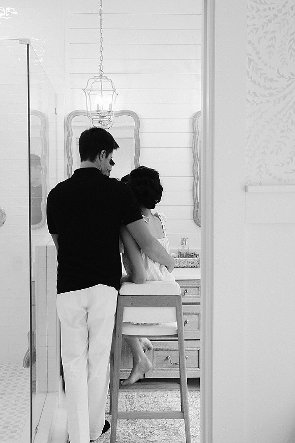 Couple portrait in black and white, bride sitting on a high chair with groom standing behind her in an intimate bathroom setting