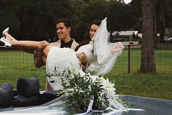 Couple portrait of groom carrying bride in veil, both laughing in a long sleeve lace wedding dress beside a convertible car on a lawn