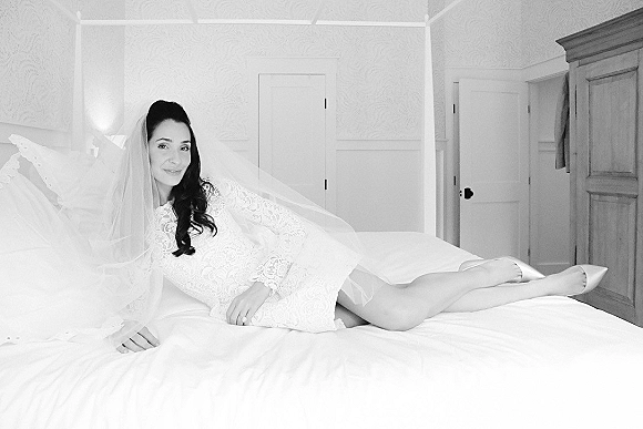 Bridal portrait in black and white of a bride reclining on a bed, veil over her face, in a lace long sleeve wedding dress in a bedroom