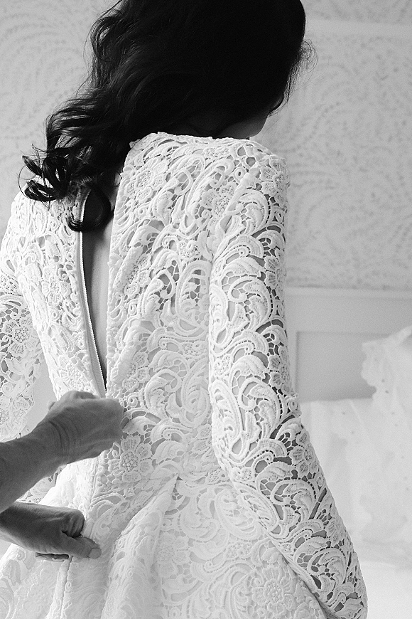 Wedding dress detail showing lace wedding dress with long sleeves and open back, highlighting the back zipper beside a bed headboard indoors