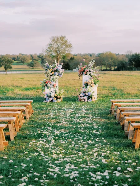 Ceremony setup for an outdoor wedding ceremony with wooden benches arranged on a grass lawn in an open field framed by trees beneath a wide sky.