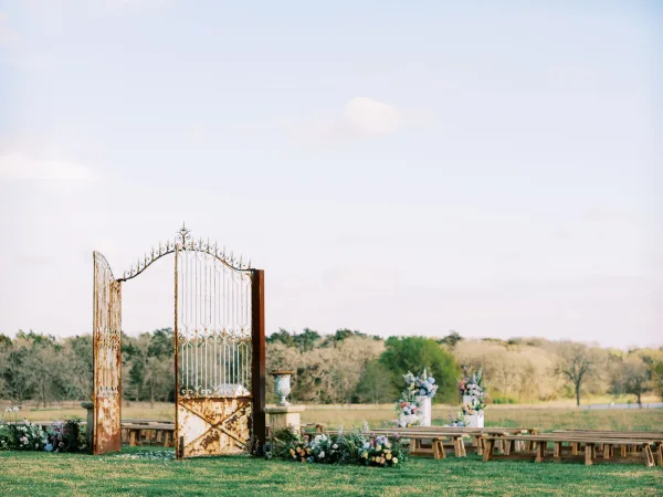 Ceremony entrance marked by an iron gate in an outdoor ceremony setup, leading to an aisle across an open field under a wide sky