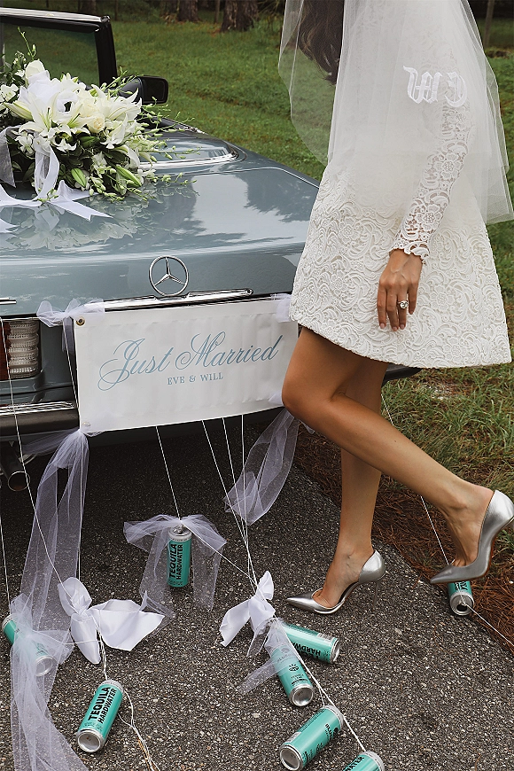 Wedding exit as bride in lace dress and veil steps into a just married car with ribbons and tin cans on a tree-lined road