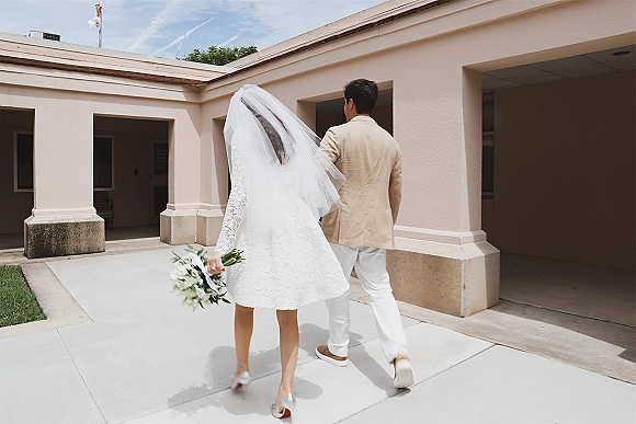 Couple portrait of bride and groom walking away, her long veil and bouquet visible, past columns of a sunny stucco courtyard walkway
