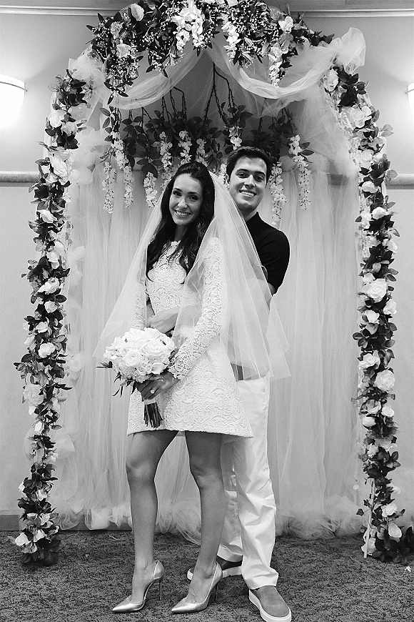 Couple portrait in a black and white wedding portrait, groom hugging bride holding bouquet before a draped floral arch and curtain backdrop