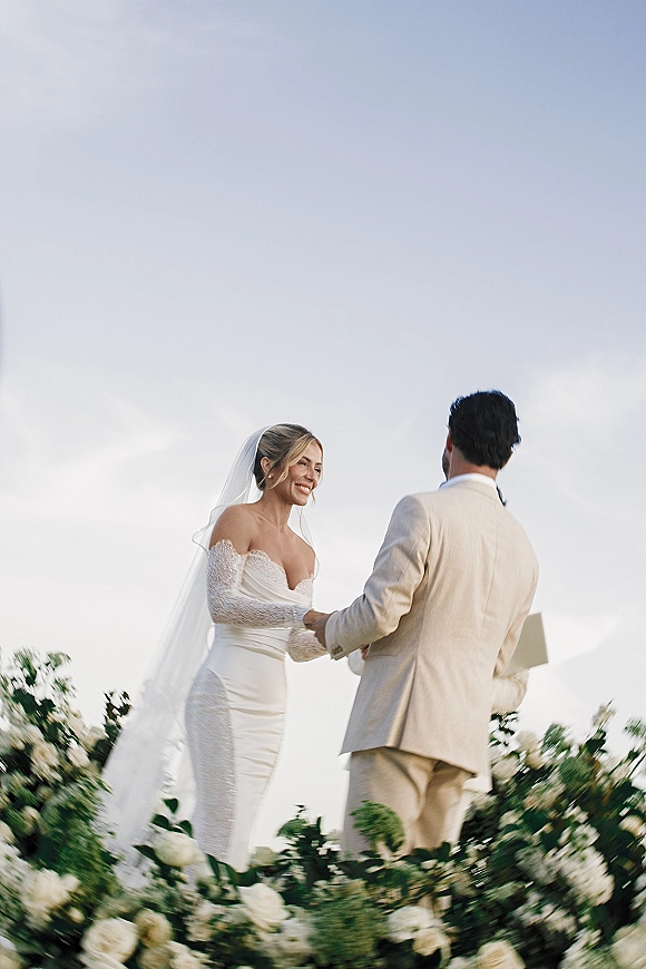 Wedding vows shared as bride and groom hold hands on an outdoor wedding ceremony platform, her veil blowing beneath open sky