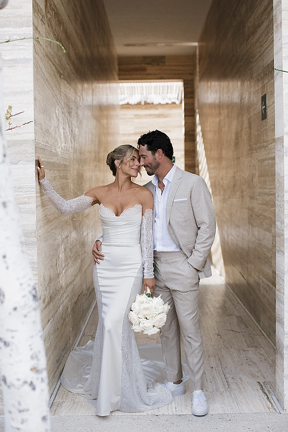 Couple portrait of bride and groom embrace with a white bouquet, forehead touch in a naturally lit stone hallway, both in sneakers