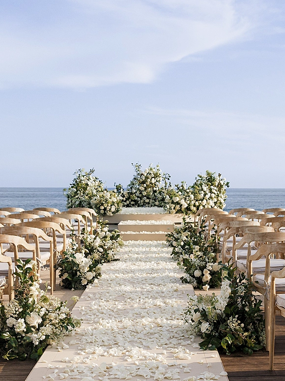 Ceremony setup for an oceanfront wedding ceremony with wood chairs, white and green florals lining a white petal aisle on a deck