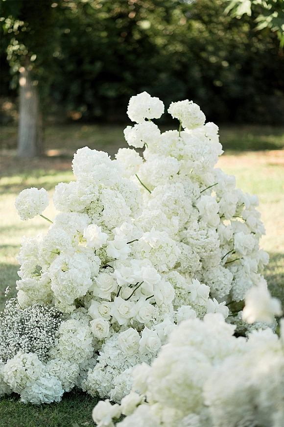 Wedding florals with a white hydrangea arrangement of white roses, baby's breath, and greenery clustered on a garden lawn with trees