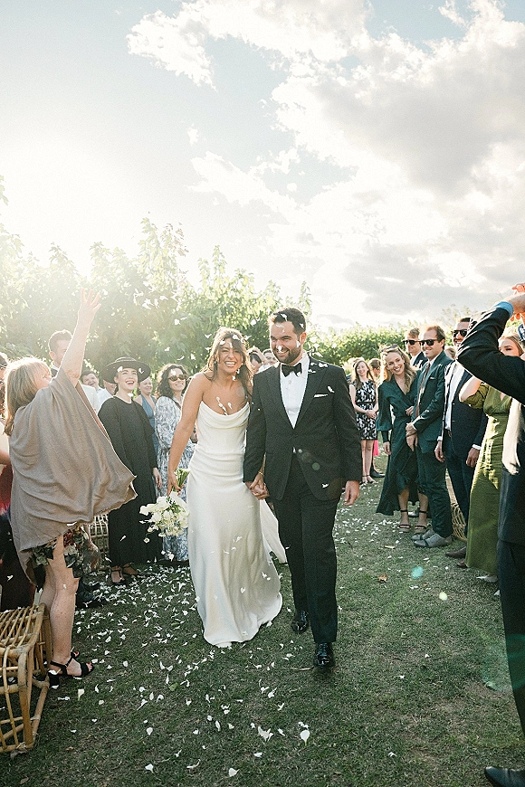 Wedding recessional as bride and groom walk hand in hand through petal confetti, bride holding white bouquet on a sunny garden lawn
