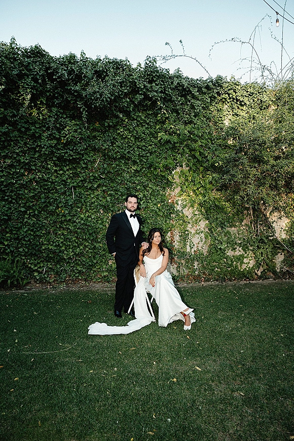 Couple portrait of bride seated on a chair in a strapless wedding dress train, groom in tuxedo beside an ivy wall with string lights