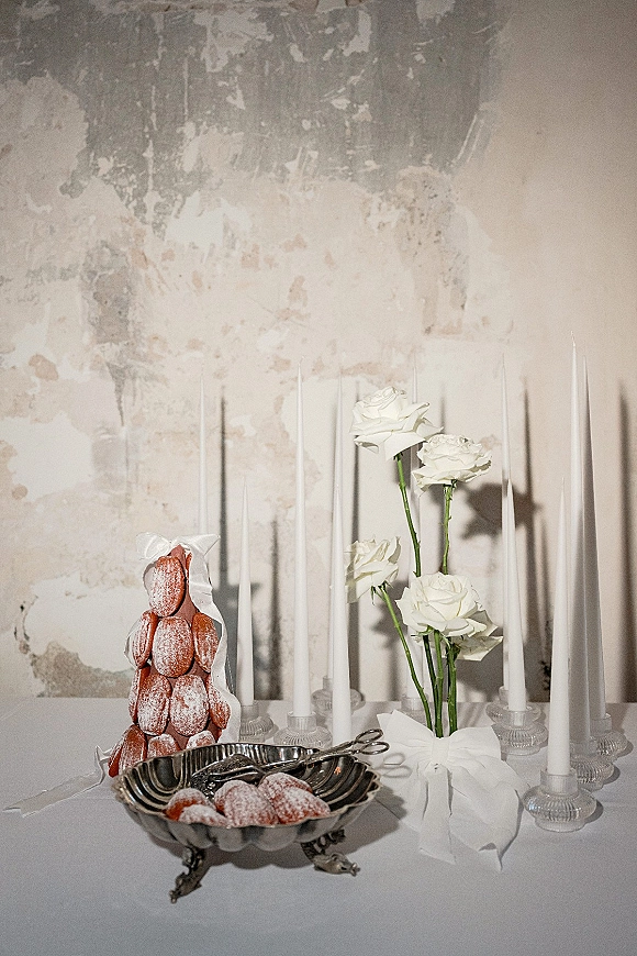 Wedding dessert table featuring a croquembouche wedding dessert with white roses, taper candles in glass holders, and pastries on a white cloth backdrop