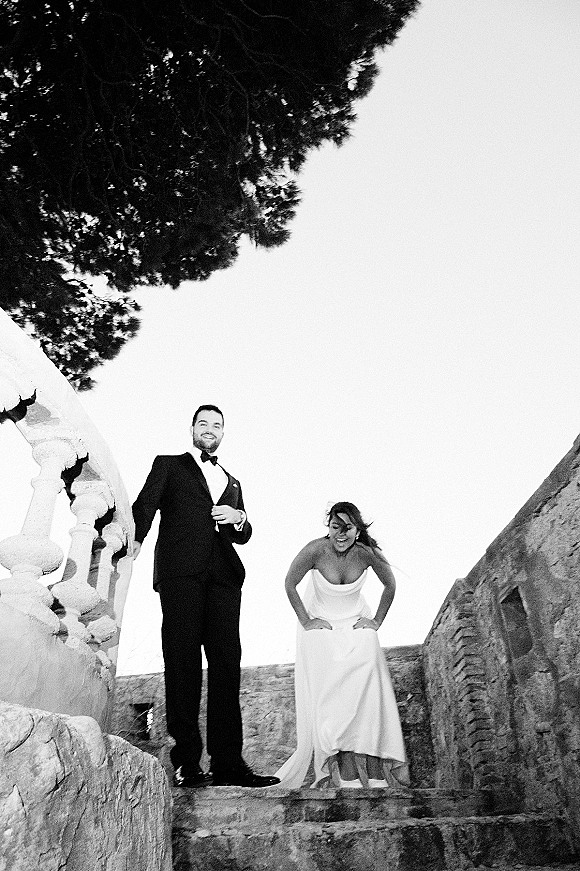 Couple portrait in a black and white wedding portrait style, bride in strapless gown and groom in tux laughing on stone stairs