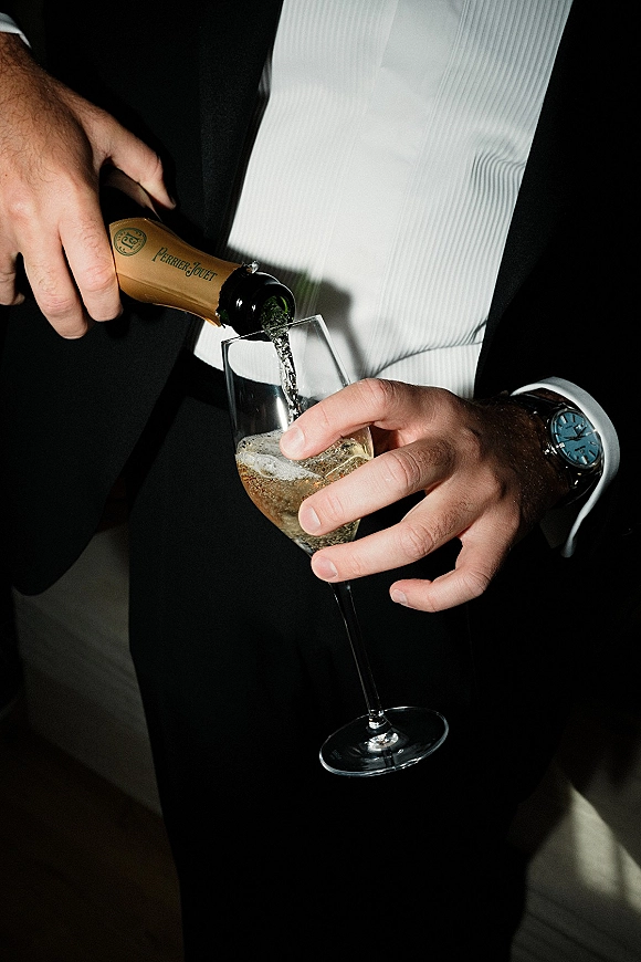 Champagne toast as a groom pours bubbly from a bottle into a flute, showing tuxedo jacket, white dress shirt and wristwatch against a dark backdrop