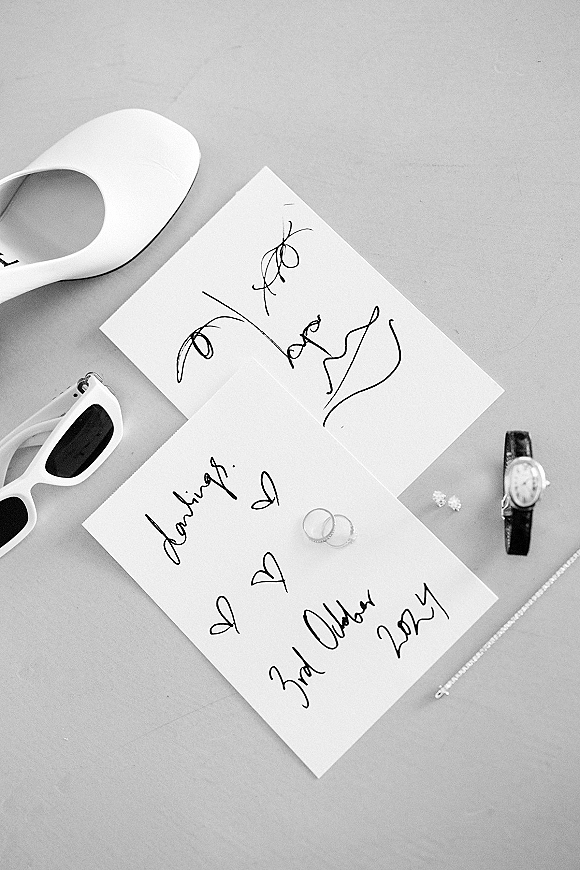 Wedding invitation suite with handwritten wedding invitation cards, rings, watch, sunglasses, necklace chain, and shoe on a neutral tabletop