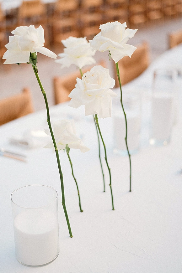 Wedding centerpieces with a single rose centerpiece in clear glass cylinder and bud vases on white linens at reception tables with wooden chairs