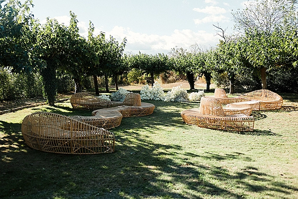 Outdoor wedding lounge with rattan wedding lounge furniture, floor cushions, and white floral arrangements on a lawn in an orchard under blue sky