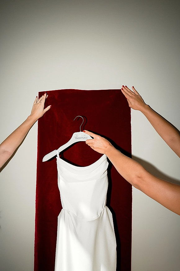 Wedding dress on a hanger held up by hands with red nail polish, a ring visible, set against a burgundy fabric backdrop and white wall