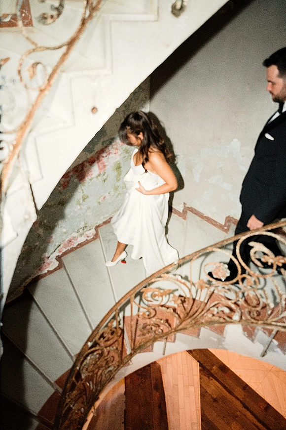 Wedding couple portrait of groom in tuxedo watching bride walking down stairs in a strapless gown, on a spiral staircase with wrought iron railing