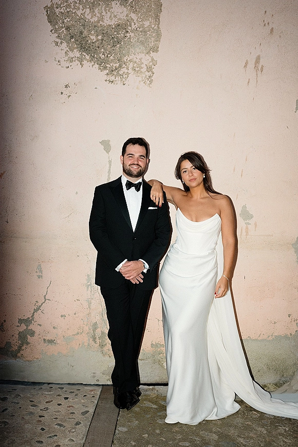 Couple portrait of bride and groom posing full length, her arm on his tuxedo, by a textured plaster wall with peeling paint and concrete floor