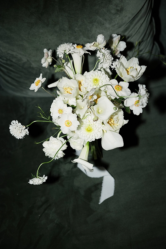Bridal bouquet of white flowers and calla lilies with greenery, wrapped in white ribbon, arranged against a dark fabric backdrop