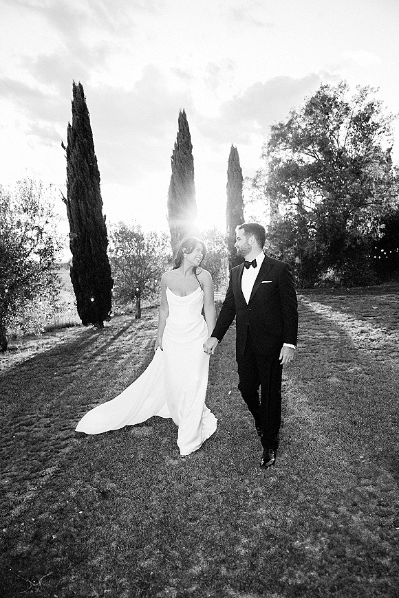 Couple portrait of bride and groom walking hand in hand, her wedding dress train flowing, on a tree-lined lawn with sun flare