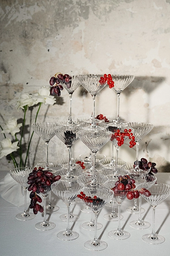 Champagne tower setup of stacked coupe glasses with grapes and red berries, white floral accents on a table against a textured plaster wall