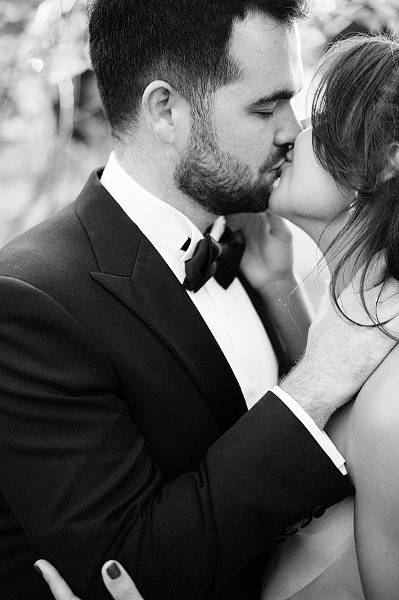 Wedding kiss portrait in black and white, bride and groom kissing close up as she holds his bearded face, tuxedo and ring in focus, greenery bokeh behind