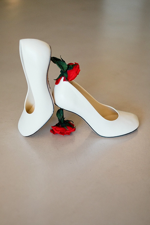 Wedding shoes styled as white bridal pumps with red roses and green leaves on a neutral floor, capturing elegant getting-ready details