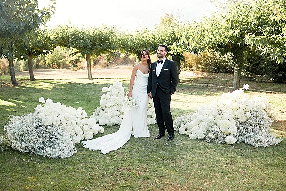 Couple portrait of bride and groom standing hand in hand, laughing in sunlit garden, bride in strapless gown with white bouquet