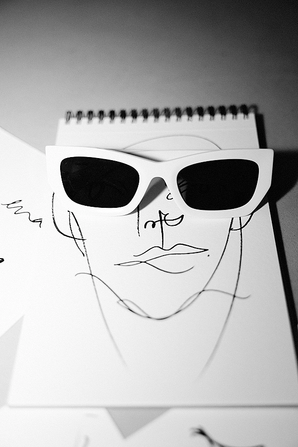 Wedding stationery flatlay with a wedding invitation suite, sunglasses accent, illustrated face card, envelope and spiral notebook on tabletop surface