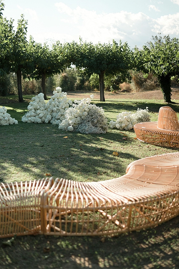 Outdoor ceremony decor with ground floral aisle decor featuring white hydrangea clusters on grass, rattan lounge seating on a sunny lawn