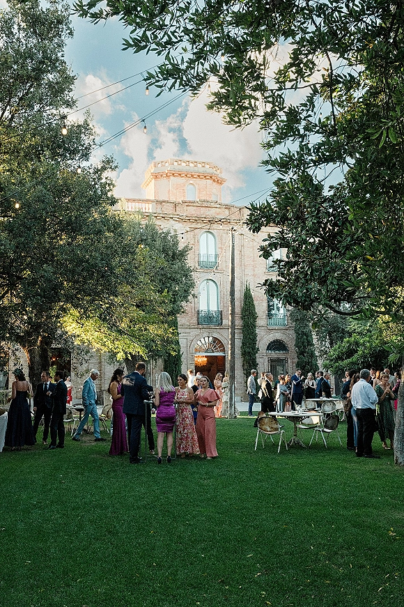 Wedding cocktail hour with guests mingling around cocktail tables under string lights on a garden lawn beside a stone building