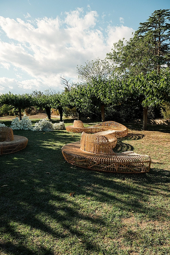 Outdoor lounge setup with wedding lounge furniture, woven rattan seating and white floral arrangements on a grassy lawn under blue sky
