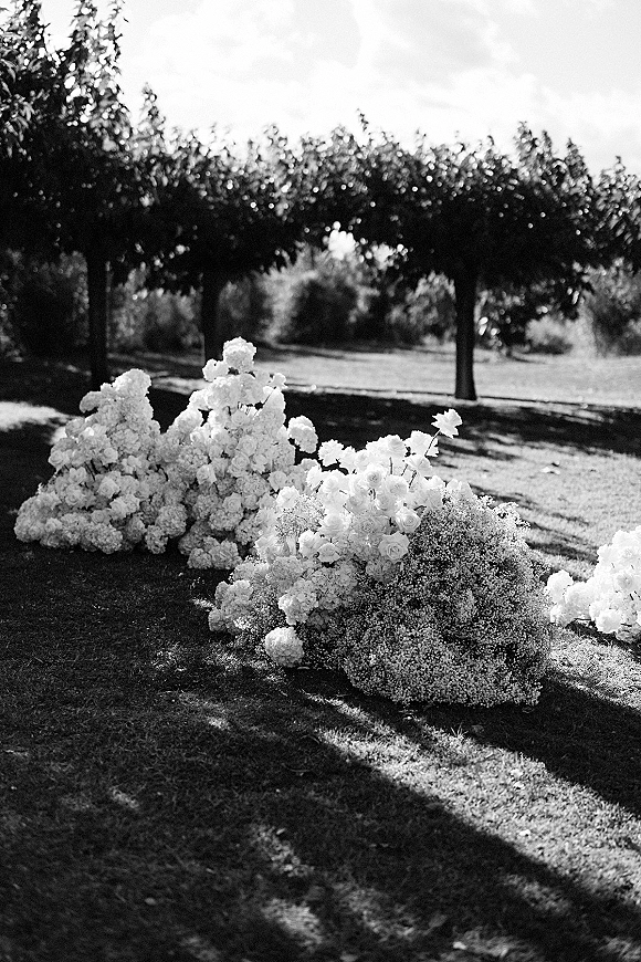 Ceremony florals with white hydrangeas and roses arranged low on the grass, creating lush clusters in a sunlit outdoor setting