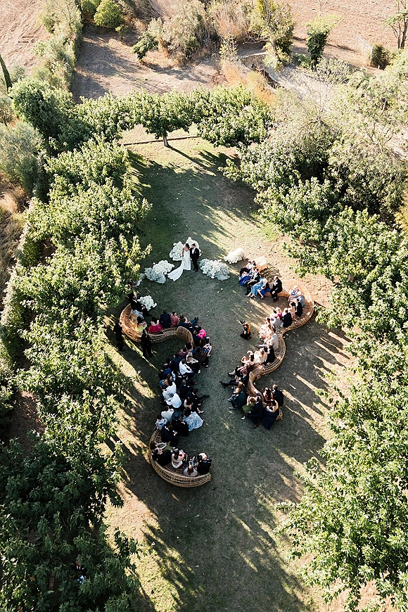 Outdoor wedding ceremony with aerial wedding ceremony view, bride and groom at altar amid white florals and curved wicker benches in a sunny tree grove
