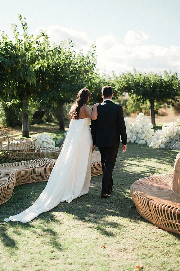 Couple portrait of bride and groom walking away, her strapless gown with long train and floral aisle arrangements on a sunny lawn
