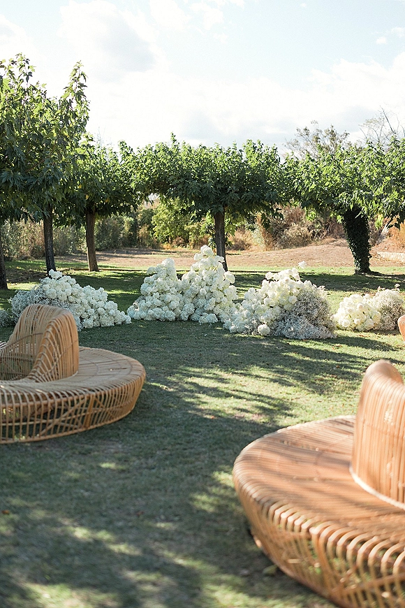 Outdoor ceremony decor with white floral ceremony arrangements lining a grounded aisle beside rattan lounge chairs on a grassy lawn under open sky