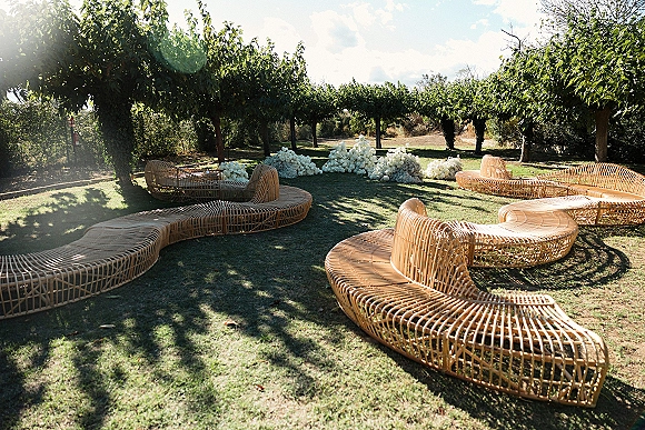 Outdoor lounge seating in a wedding lounge area with curved rattan sofas and white floral arrangements on a sunny lawn with trees and hedges