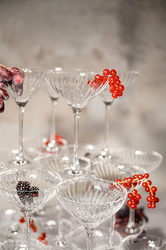 Champagne tower of coupe glasses stacked on a tabletop, garnished with red berries, blackberries, and grapes against a neutral wall