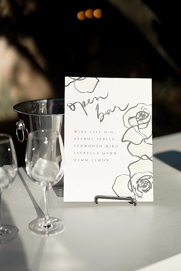 Wedding bar menu and open bar sign in calligraphy on a menu card with floral illustration, on a tabletop beside wine glasses and ice bucket