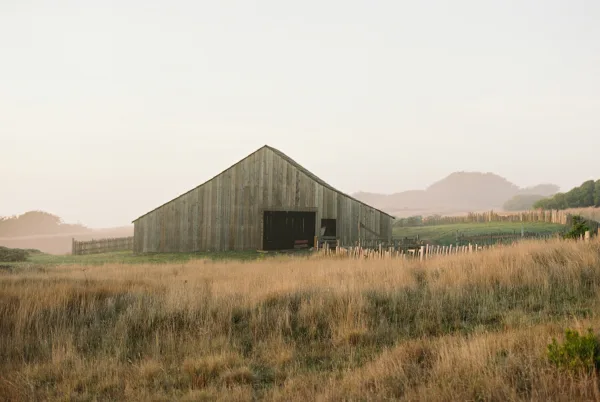 Rustic barn venue with weathered wood barn doors and fence posts beside tall grass, framed by foggy rolling hills and trees