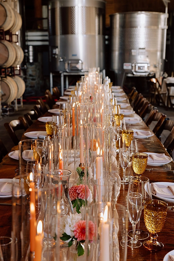 Reception tablescape on a long wedding table with taper candles, pink and white bud vases, amber goblets, in an industrial barrel room