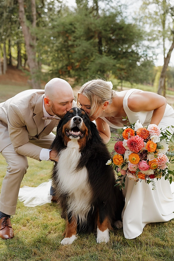 Couple with dog sharing a kiss with their pet as the bride holds a colorful bouquet, standing on a grassy lawn with trees behind