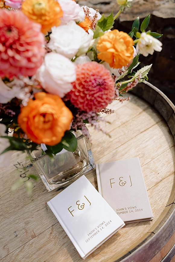 Wedding vow books with personalized vow books in gold foil lettering and monogram initials beside a bright floral centerpiece on a wood barrel tabletop