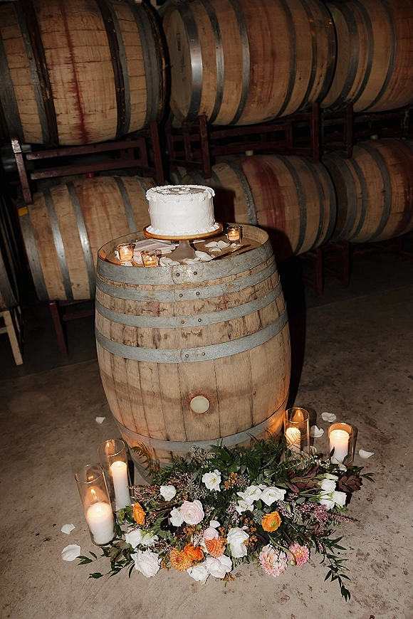 Wedding cake table with a white frosted cake on a barrel cake table, surrounded by candlelight, greenery garland, and rose petals in a winery room