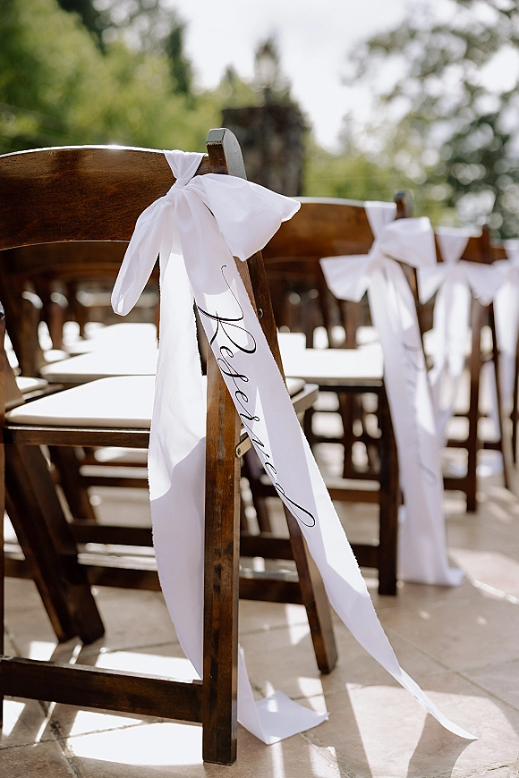 Ceremony chair decor with wedding chair bows on wood folding chairs, script reserved signs, on a sunlit outdoor patio with greenery